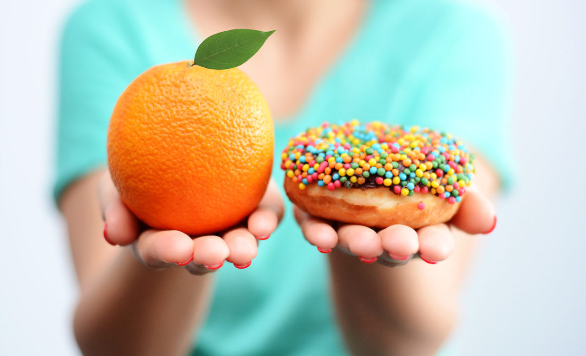 Young woman holding in hands an orange and a tasty multicolored donut, choosing healthy food or dessert Odchudzanie